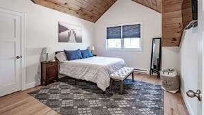 Ein Schlafzimmer mit schrägen Wänden, einem Bett mit weißer Tagesdecke und blauen Kissen, einem gemusterten Teppich, einer Bank, einem Fenster mit Jalousien und einer Tür.