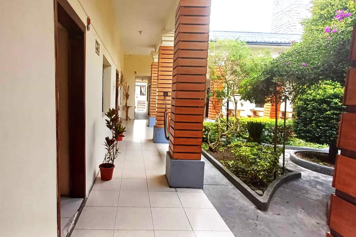 A corridor with tiled flooring, wooden pillars, potted plants, and a view of a garden with trees and shrubs.
