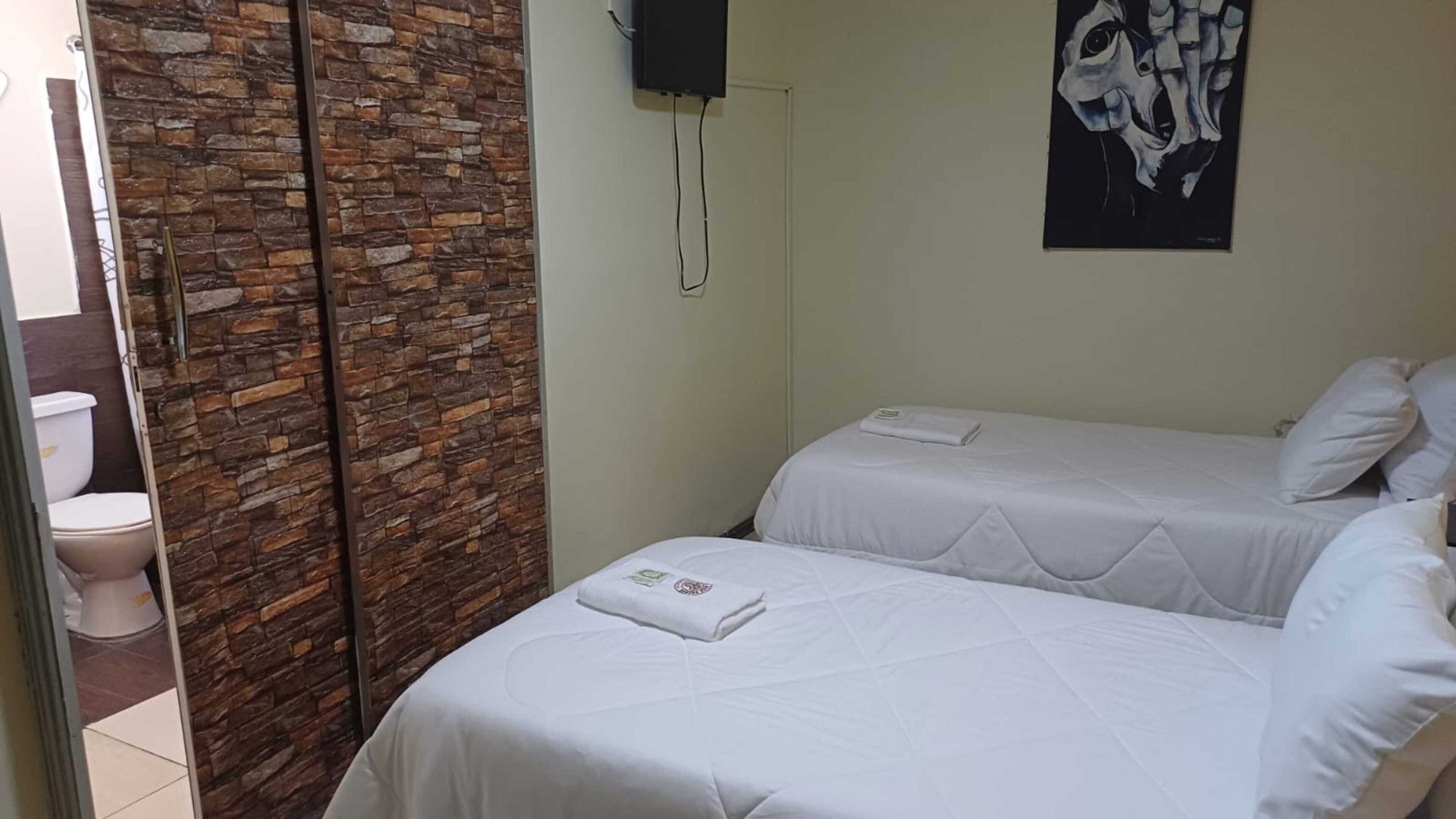 Une chambre d’hôtel avec deux lits, un mur en pierre, un tableau et des toilettes visibles à travers une porte ouverte.