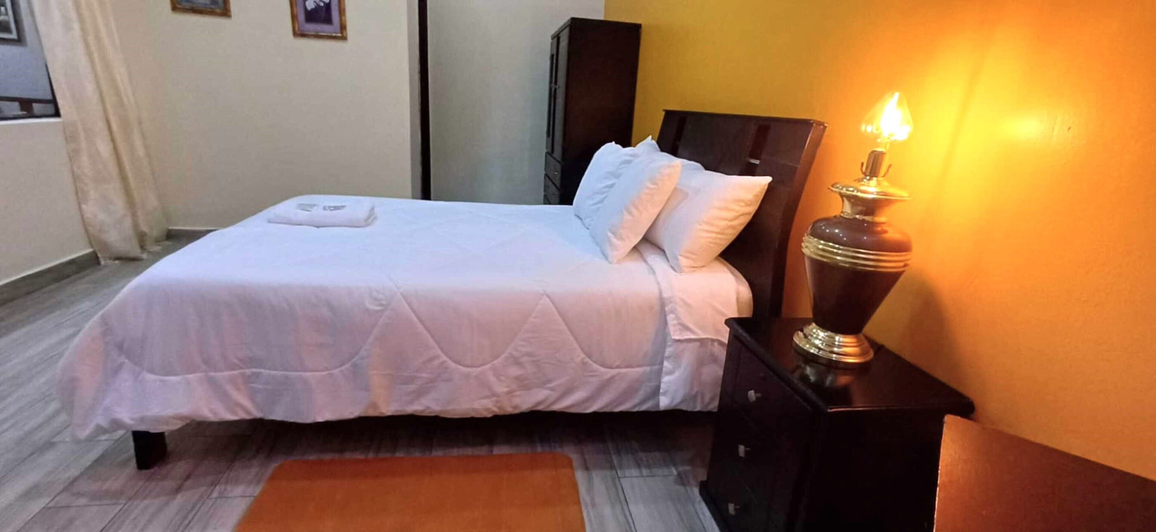 A neatly made bed with a white duvet and pillows, a bedside table with a lamp, and a wooden nightstand.