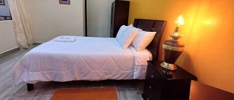 Una cama bien hecha con un edredón blanco y almohadas, una mesita de noche con una lámpara y un mueble auxiliar de madera.
