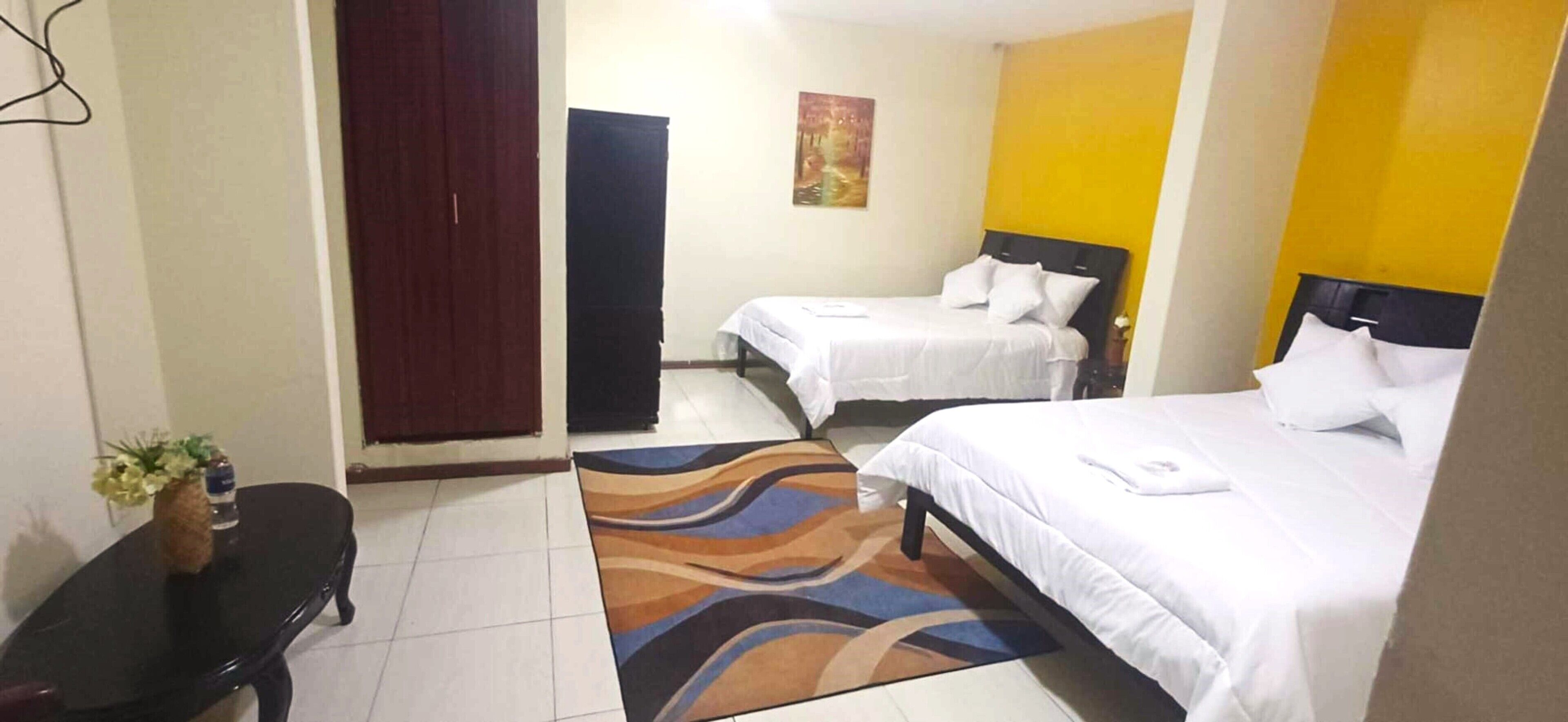 A hotel room with two beds, a small table with a vase, and a colorful rug.