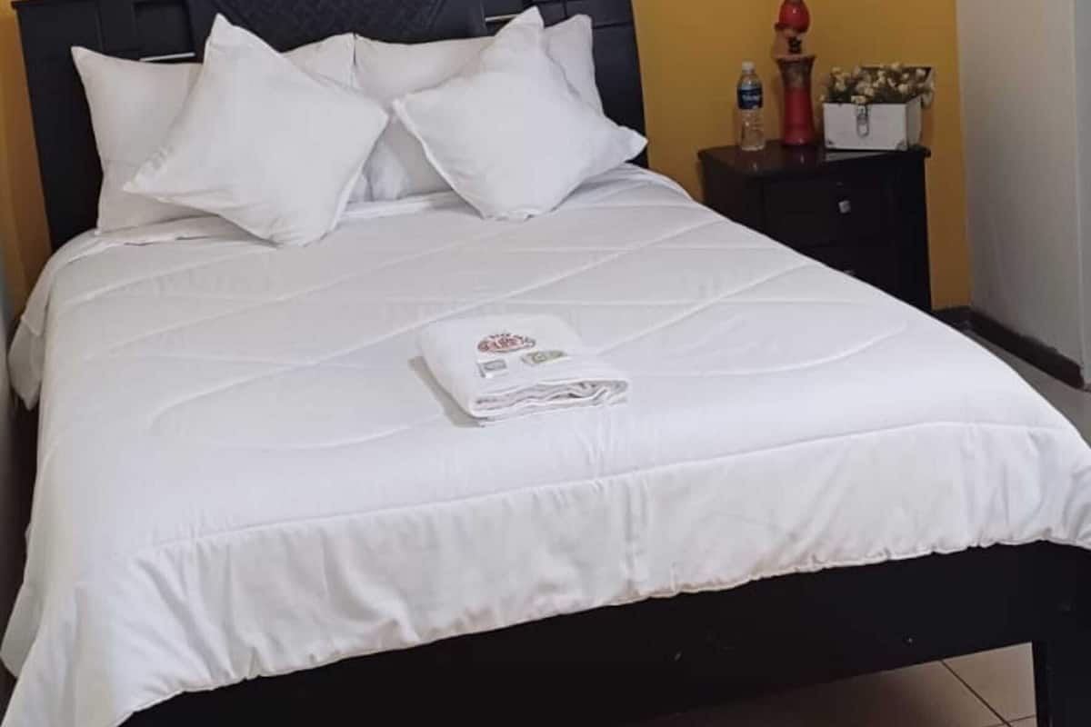 A neatly made bed with white bedding and pillows, a brown floor mat, and a nightstand with a water bottle and decorative items.