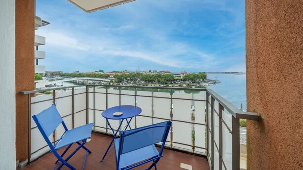 A balcony with two blue chairs and a small table, overlooking a waterfront with buildings and a bridge. - GuestHost - Relais Rosanna Stunning Lagoon-View Apartment (Grado)