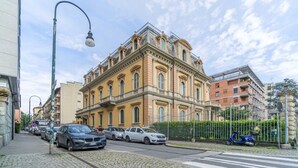 Ein historisches Gebäude im klassischen Stil, eine Kopfsteinpflasterstraße und geparkte Autos.