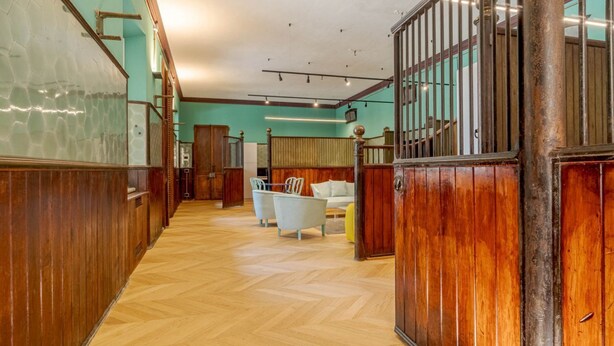 Eine Hotelhalle mit Holzwandverkleidung, einem Fischgrätparkett, einer Treppe und einer Sitzecke mit Stühlen und einer Couch.