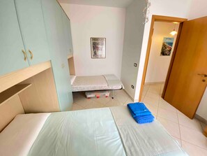 Une petite pièce comprenant un lit, un bureau pliant, une étagère fixée au mur et une porte donnant sur une autre pièce.