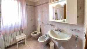 Une salle de bain comprenant des toilettes, un bidet, un lavabo, un miroir et une fenêtre avec des rideaux légers.