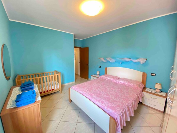 Une chambre à coucher avec un berceau, un lit recouvert d’une couverture rose, une table de chevet avec une lampe et une commode en bois.