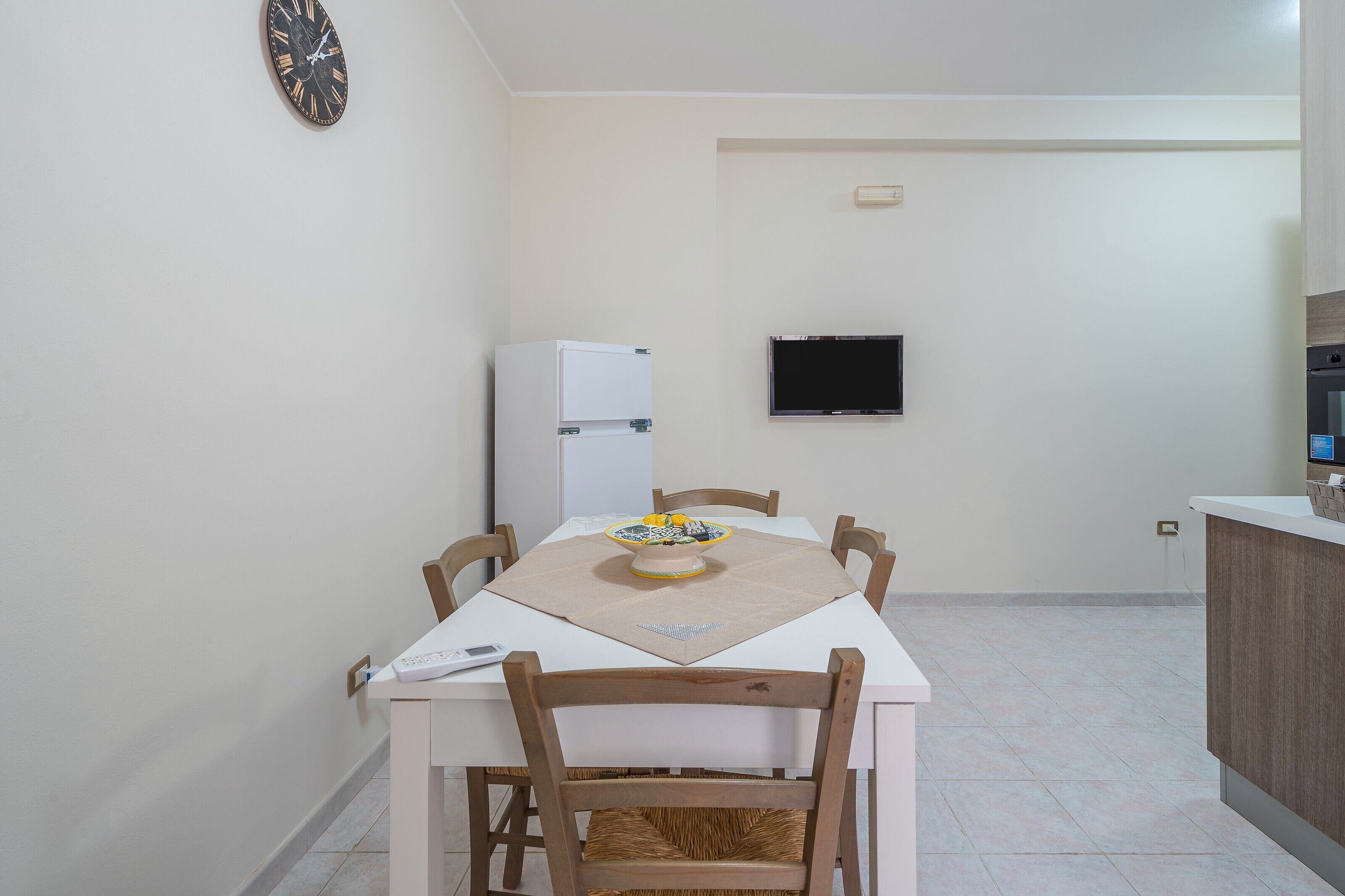 A dining area with a table, chairs, a decorative bowl, a clock, a TV, and a refrigerator.