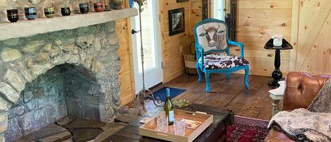 Interior rústico de una cabaña con chimenea de piedra, paredes de madera, un sillón azul, sofá de cuero y una mesa de centro de madera con una botella y vasos.