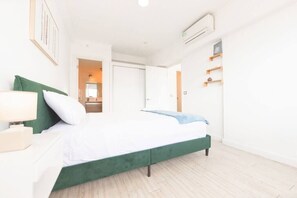 Un dormitorio moderno con una cama tapizada en verde, ropa de cama blanca, una mesita de noche con lámpara y un aire acondicionado instalado en la pared.