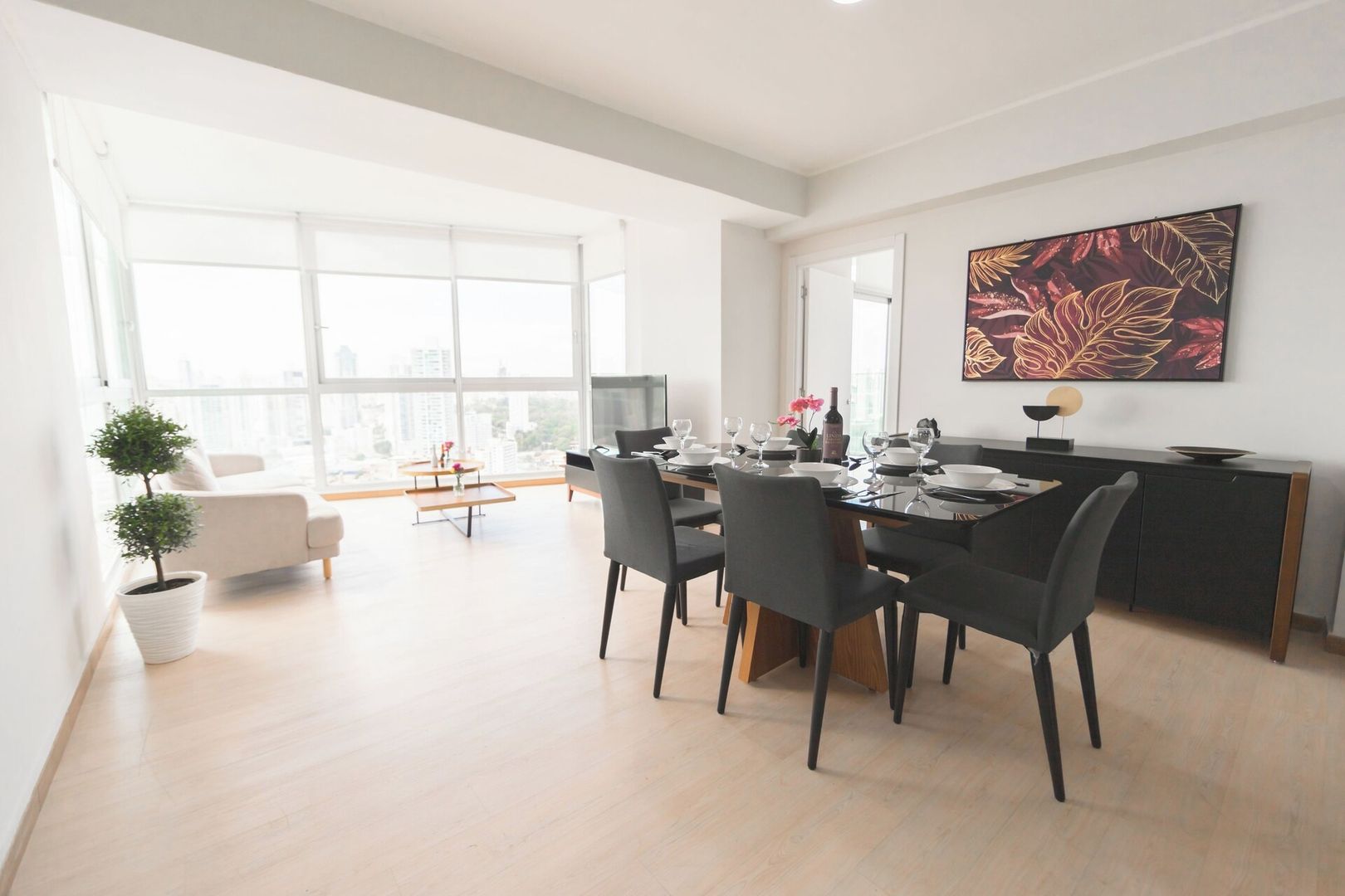 Un'area pranzo moderna con un tavolo grande apparecchiato per quattro persone, vista sulla città attraverso ampie finestre e un quadro contemporaneo appeso al muro.