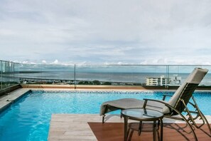 A rooftop pool with a glass balustrade, two lounge chairs, and a view of the ocean and distant buildings.