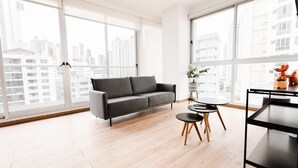 A modern living room with a grey sofa, a small round table, and a black coffee table. Large windows offer a view of tall buildings.