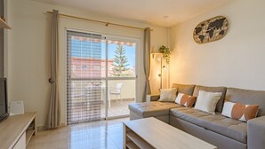 A modern living room with a sofa, a coffee table, and a TV. There is a sliding glass door leading to an outdoor area with a chair and a plant.