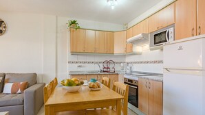 A compact kitchen with wooden cabinets, a microwave, and a refrigerator. There is a dining table with chairs and a bowl of fruit.