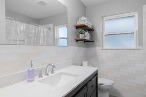 Une salle de bain avec un lavabo blanc, un miroir, un rideau de douche, des toilettes et des étagères contenant des serviettes et des articles de toilette.