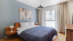 Een moderne slaapkamer met een houten hoofdbord, een groot bed met een blauwe deken, twee abstracte schilderijen aan de muur, een hangende pendantlamp en uitzicht buiten door een raam met gordijnen.