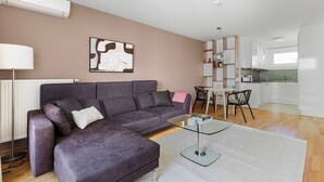 Een moderne woonkamer met een grijze hoekbank, een glazen salontafel en een eethoek met een witte tafel en stoelen.