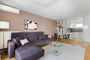 Un salon moderne avec un canapé d’angle gris, une table basse en verre et un coin repas comprenant une table et des chaises blanches.