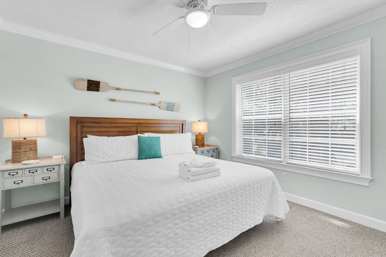 A neatly made bed with a wooden headboard, two bedside lamps, a window with blinds, and a ceiling fan.