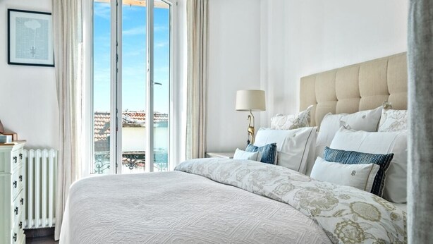A bedroom with a large bed, a bedside table with a lamp, and a view of the outside. - Upstays Banane Center 80m² Apartment (Cannes)