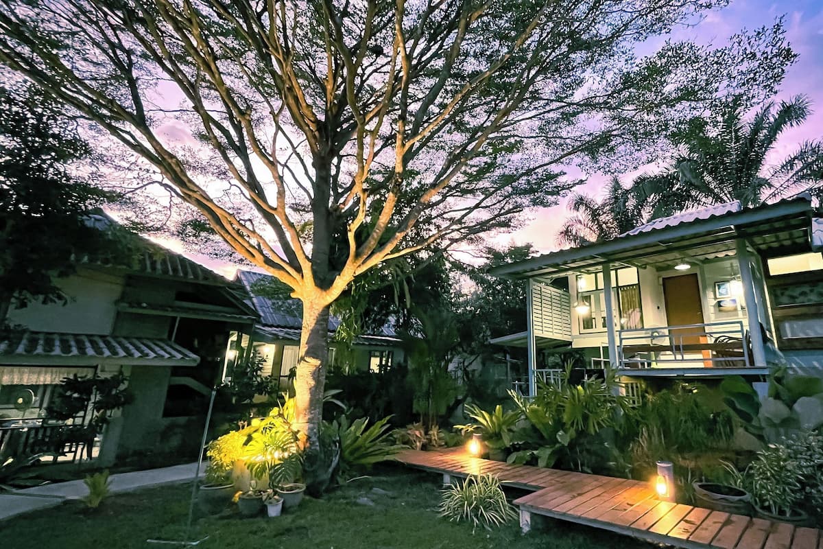 A two-story house with a large tree in the front yard, a wooden pathway, and a small porch with a railing.