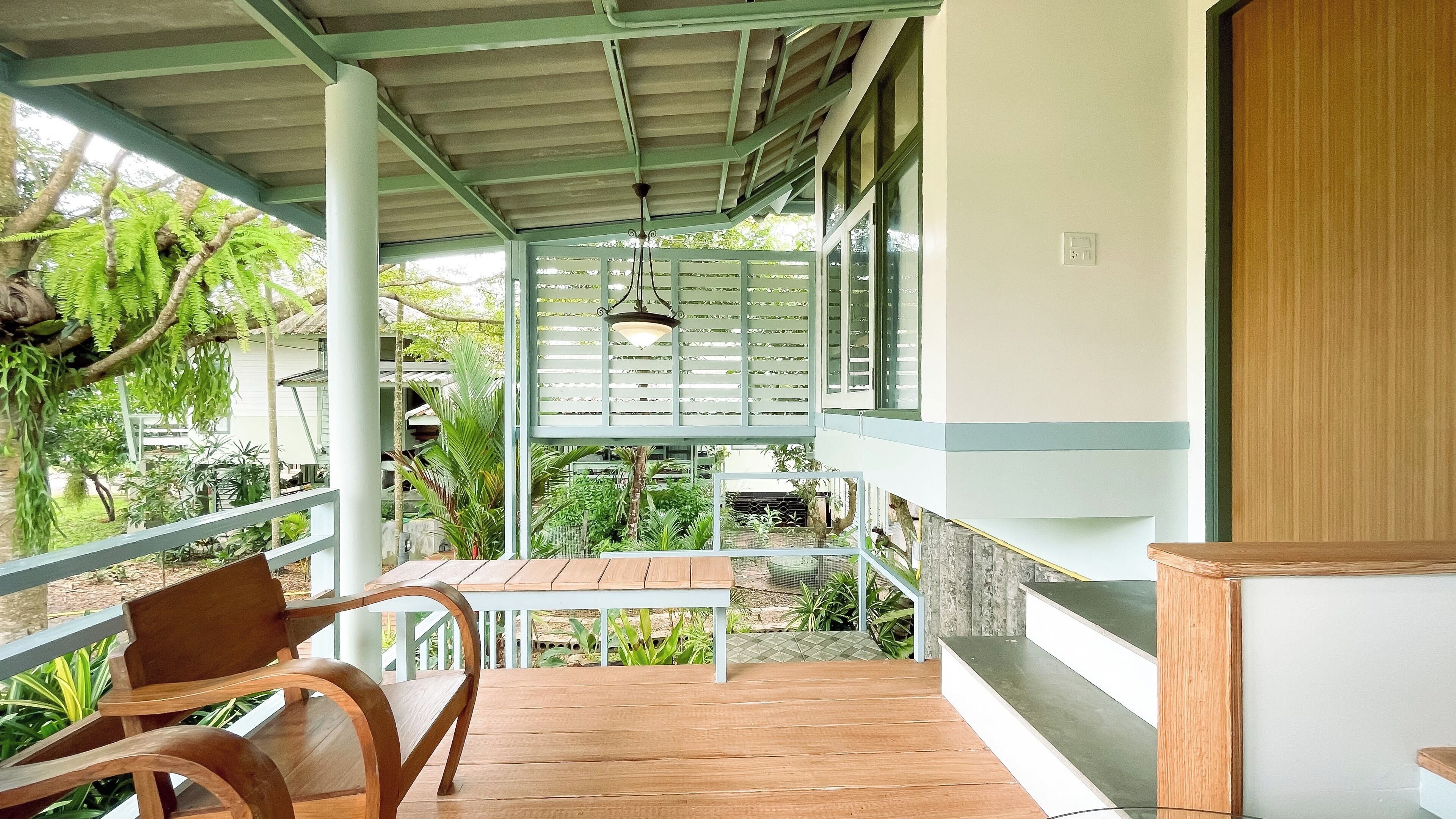 A covered outdoor area with a wooden table and chairs, a hanging light fixture, and a view of a garden with various plants and a small pond.