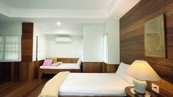 A hotel room with a bed, a small sofa, a bedside table with a lamp, wooden paneling, a window with curtains, and an air conditioning unit.