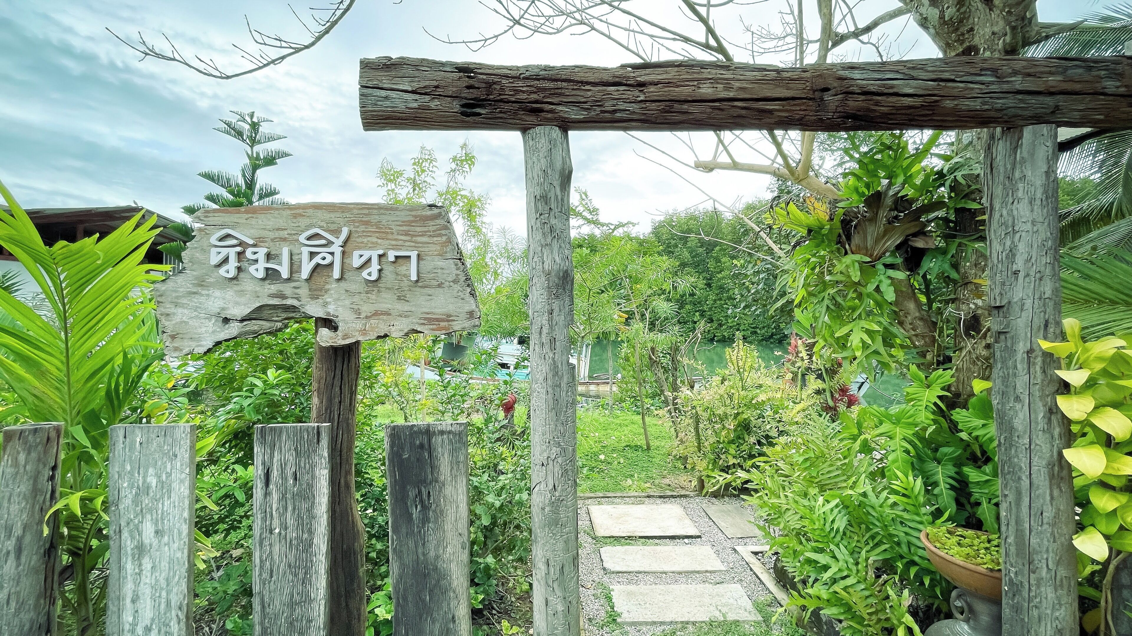 A wooden archway with a sign, surrounded by lush greenery and a pathway leading into a garden.