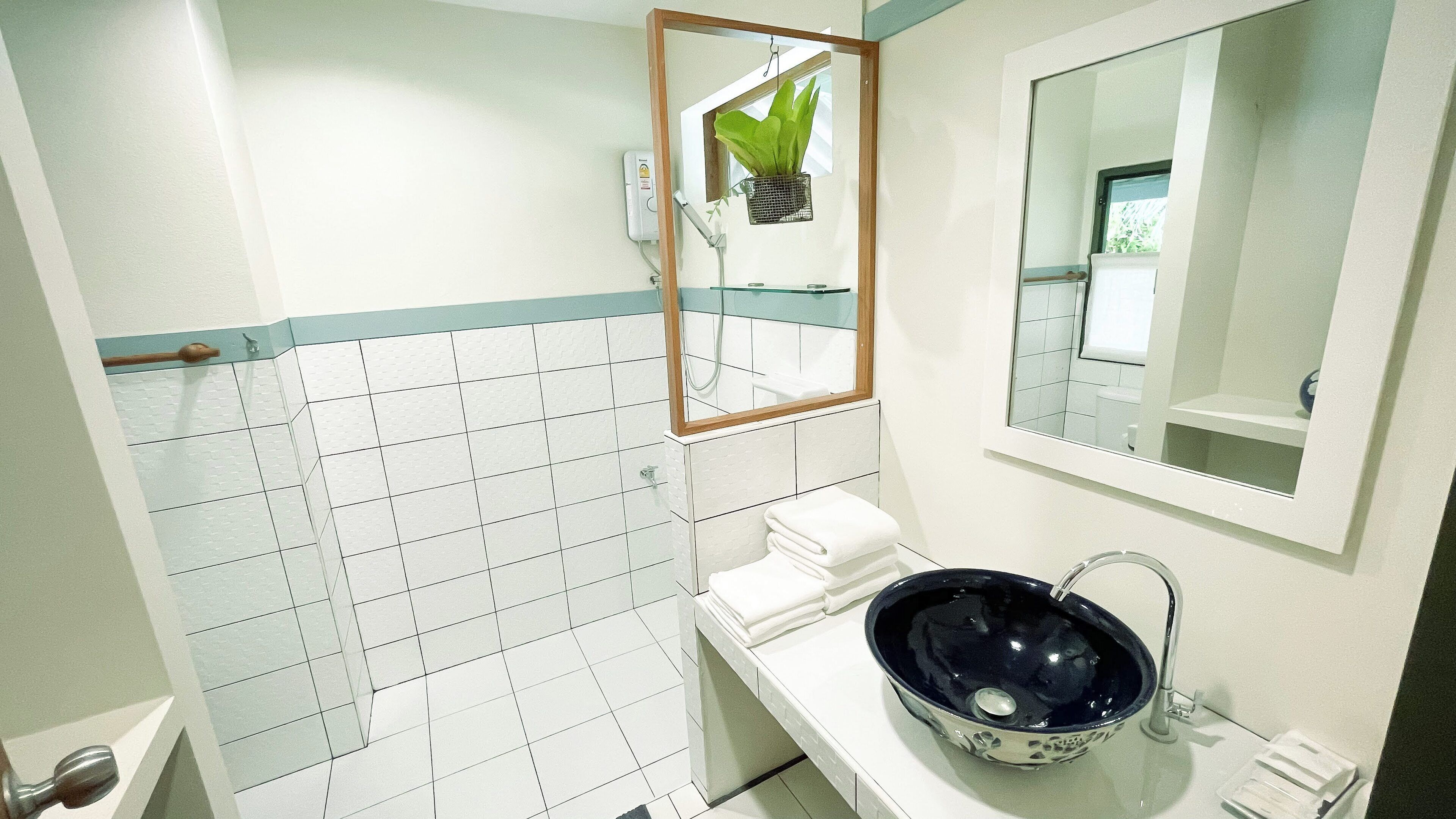 Baño con paredes de azulejos blancos, una zona de ducha con azulejos azules, un lavabo blanco con lavamanos azul y un espejo con marco de madera.