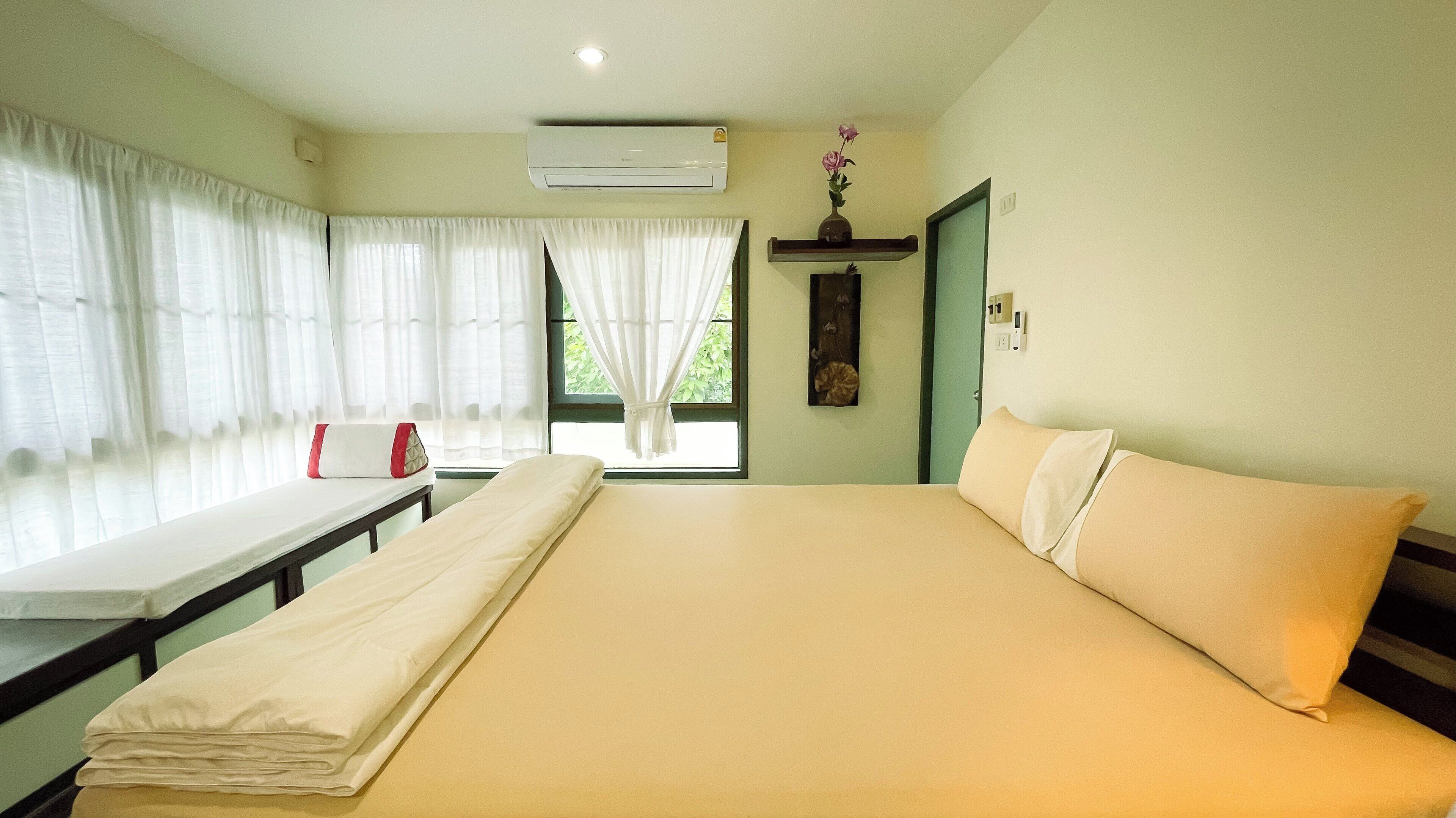 A hotel room with a bed, a window with curtains, an air conditioning unit, and a small shelf with a vase.
