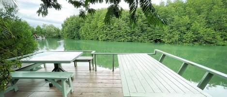 Una terraza de madera con mesas de picnic da a un río tranquilo, rodeada de abundante vegetación.