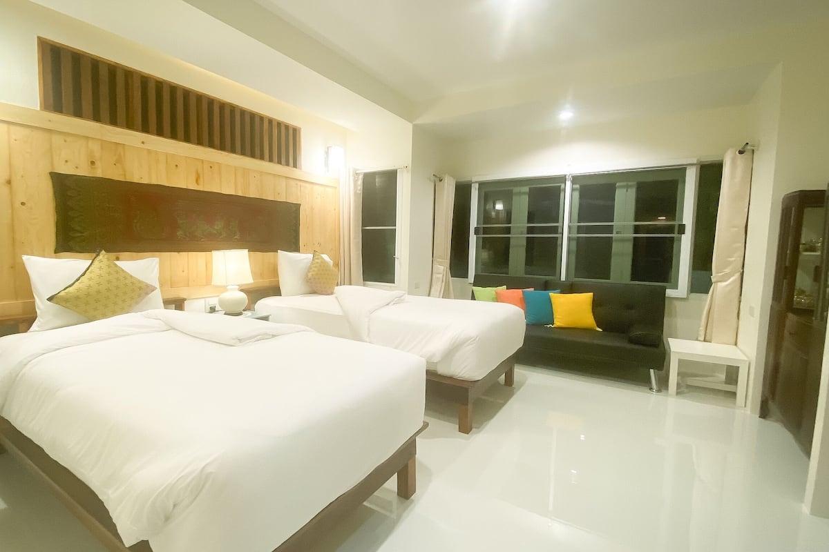 A hotel room with two beds, a wooden headboard, a sofa with colorful cushions, a small table, and a window with curtains.