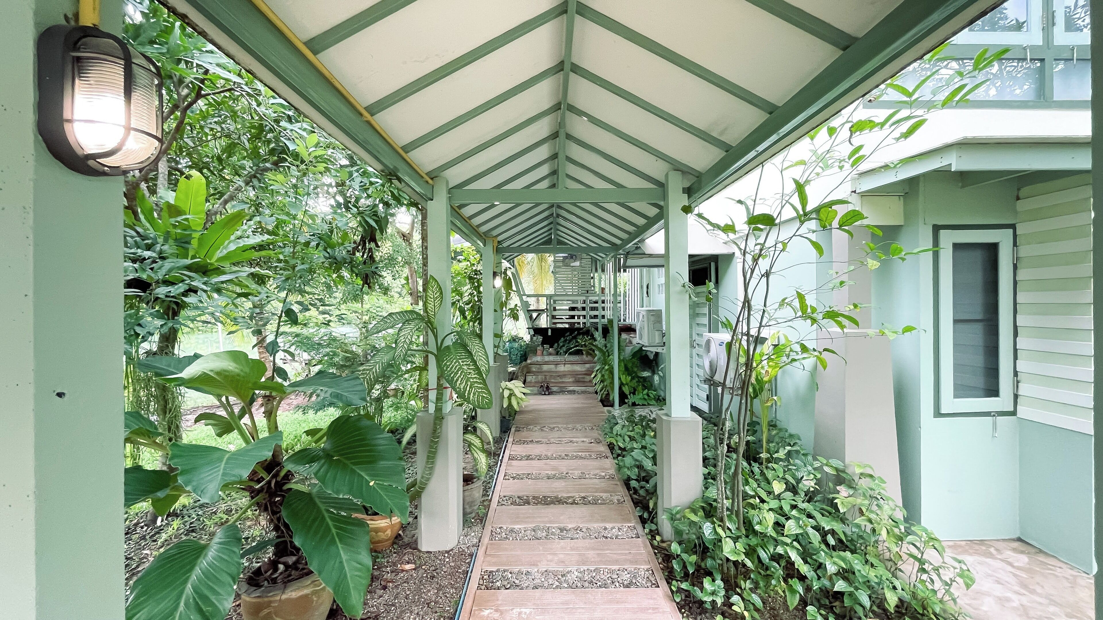 Une allée extérieure abritée, dotée d’un plancher en bois, et bordée d’une végétation luxuriante et de plantes en pot.
