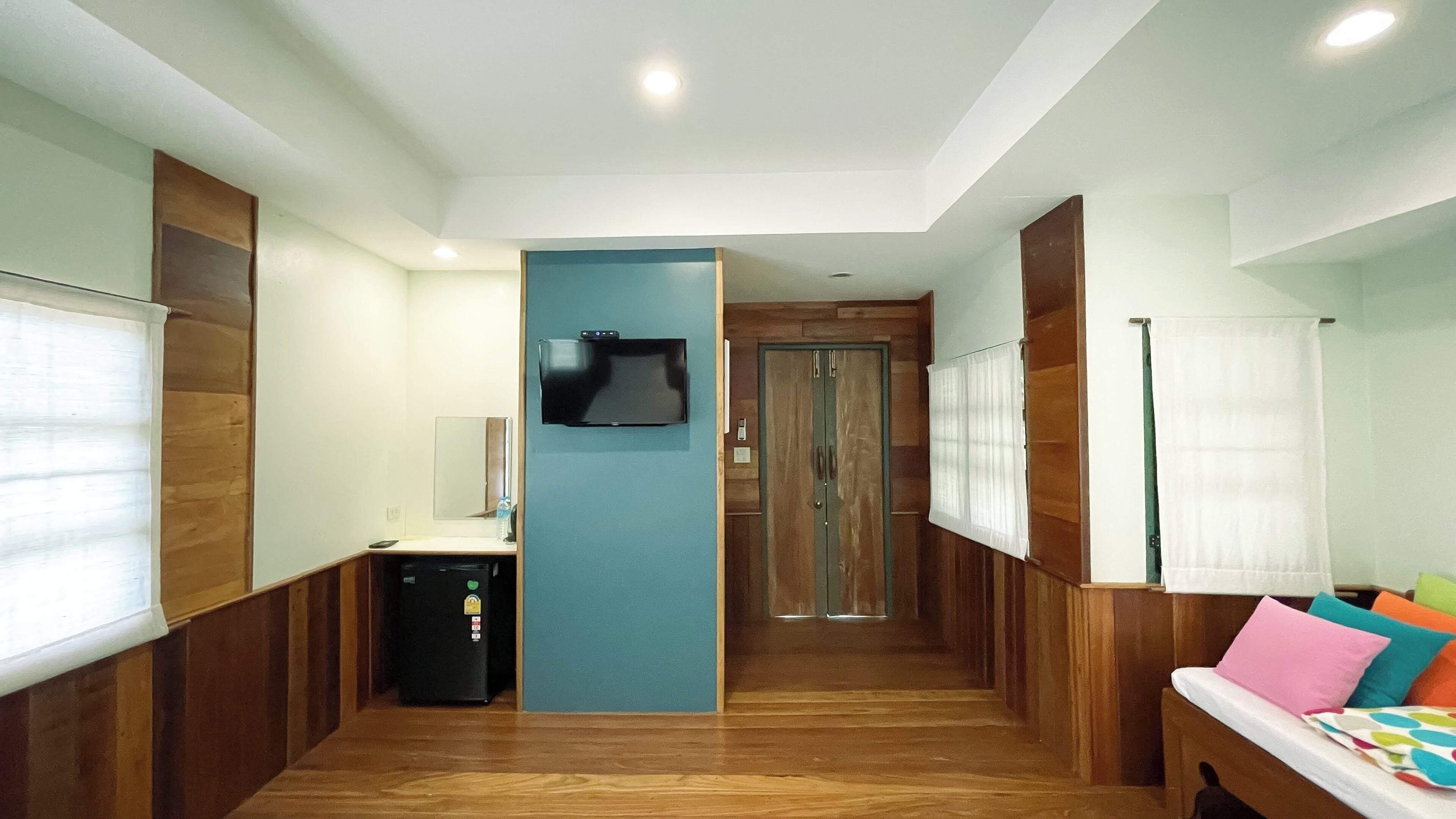 Una camera d'albergo con pavimento in legno, un letto con cuscini colorati, una televisione montata a parete e una porta che dà su un'altra stanza.
