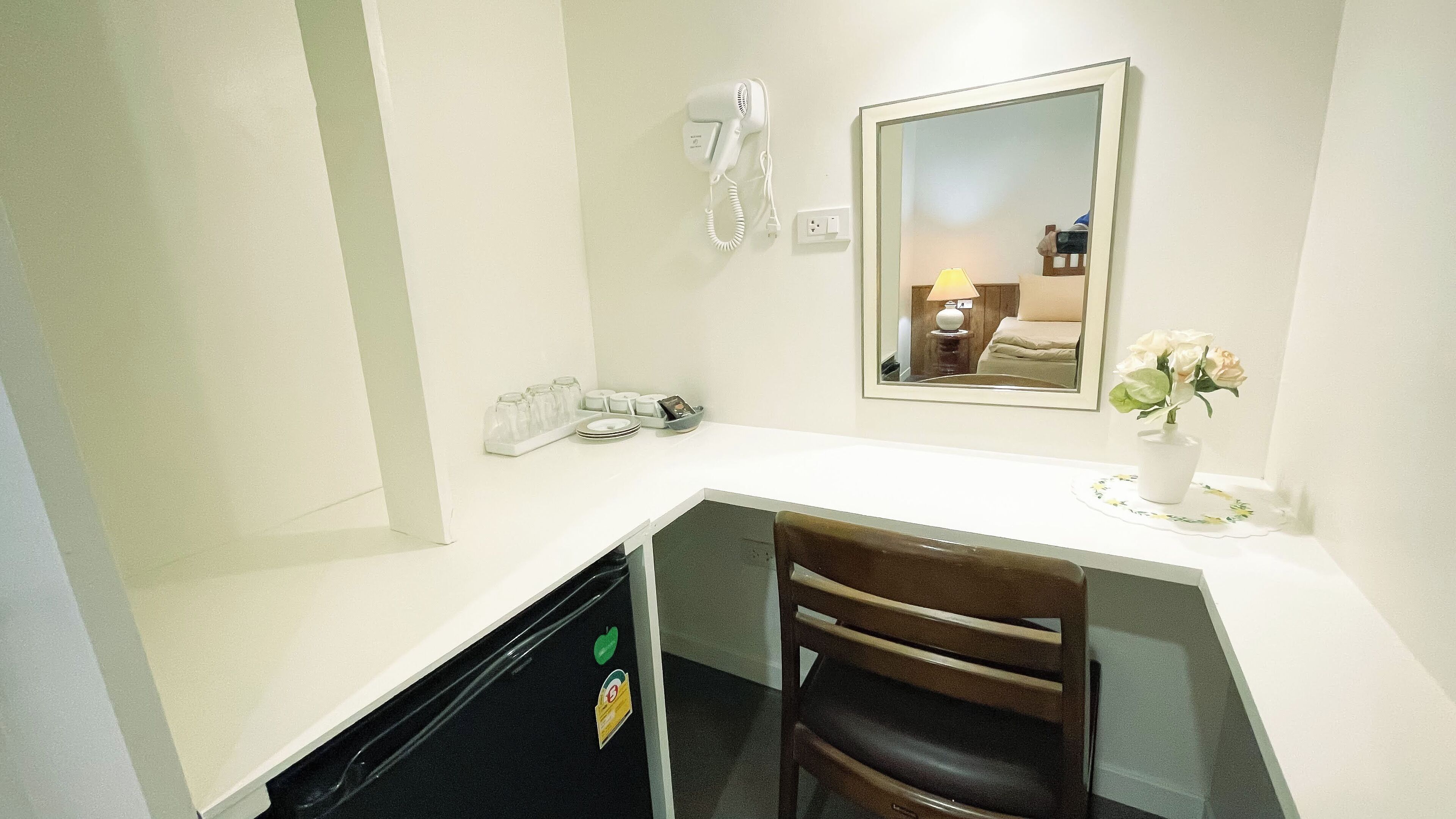 A compact hotel room with a white desk, a chair, a hairdryer, a mirror, and a small vase with flowers.