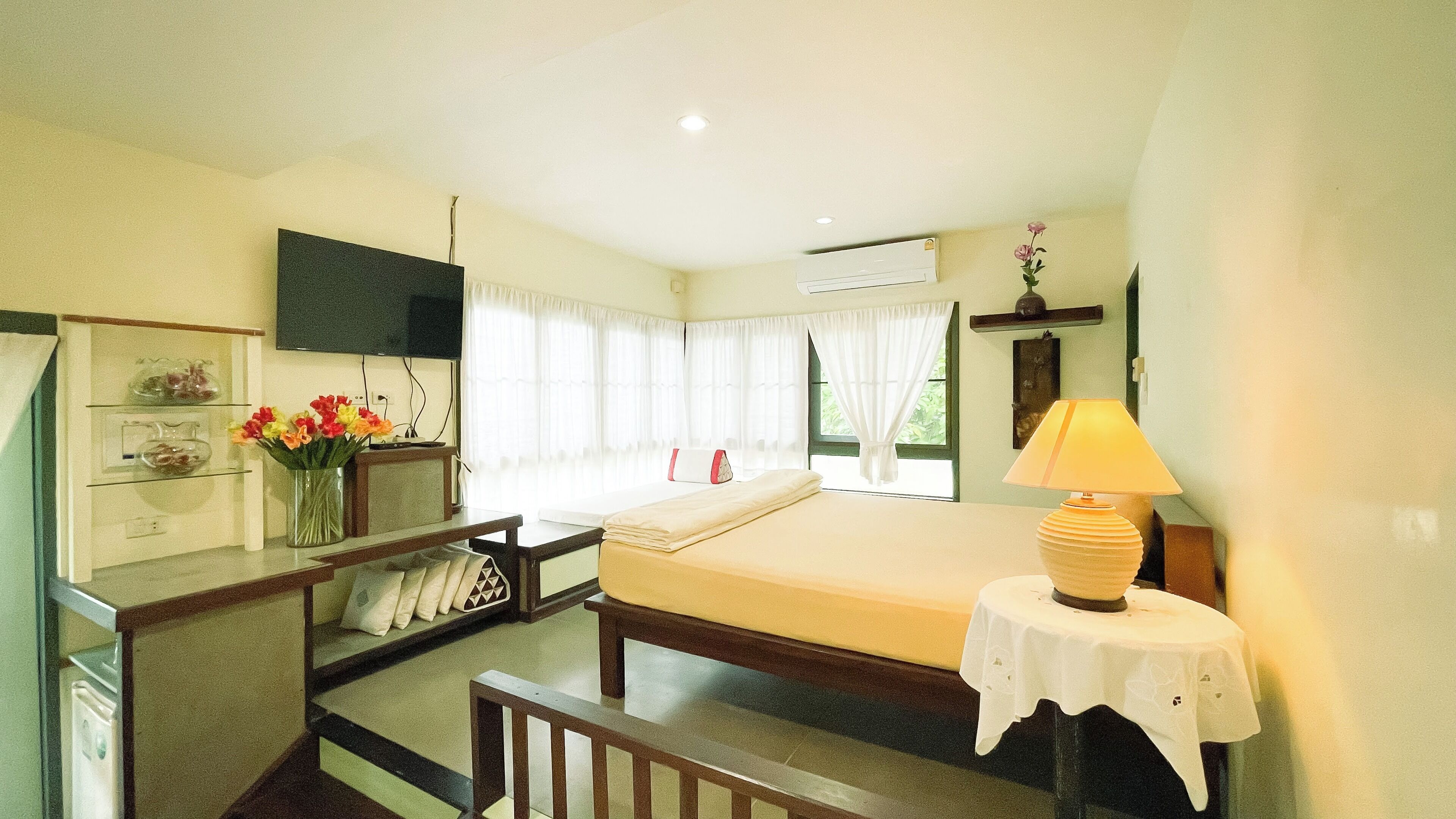 A hotel room with a bed, a TV, a desk with a vase of flowers, and a window with curtains.