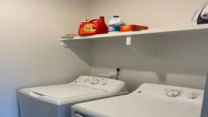 A laundry area with two front-loading washing machines and a shelf above containing detergent and other cleaning supplies.