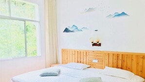 Ein Schlafzimmer mit einem hölzernen Kopfteil, einem Bett mit weißen Leinen, zwei Kissen, einem Fenster mit Vorhängen und einer Wanddekoration von Bergen und Vögeln.
