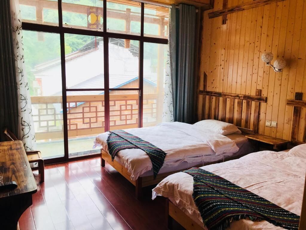 A room with two beds, wooden walls, a large window with a view of a traditional building, and a small table with a lamp.