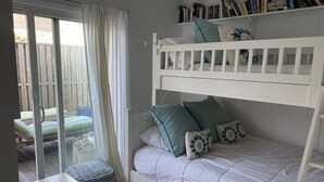 Een stapelbed met een wit frame, een onderste bed met een witte deken en een bovenbed met een wit hoofdbord. Daarboven bevindt zich een boekenplank met boeken en decoratieve objecten. Een schuifdeur leidt naar een buitenruimte met een houten terras en een stoel.