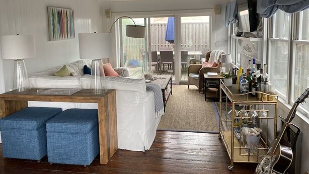 Een knus ingerichte kamer met een bed, een houten bijzettafeltje, een blauwe poef, een gitaar en een trolley met flessen.