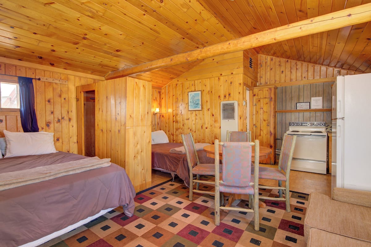 A cabin interior with a wooden interior, a bed, a small table with chairs, a kitchenette, and a window with curtains.