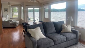Ett rymligt vardagsrum med en grå soffa, trägolv och stora fönster med utsikt över en sjö.