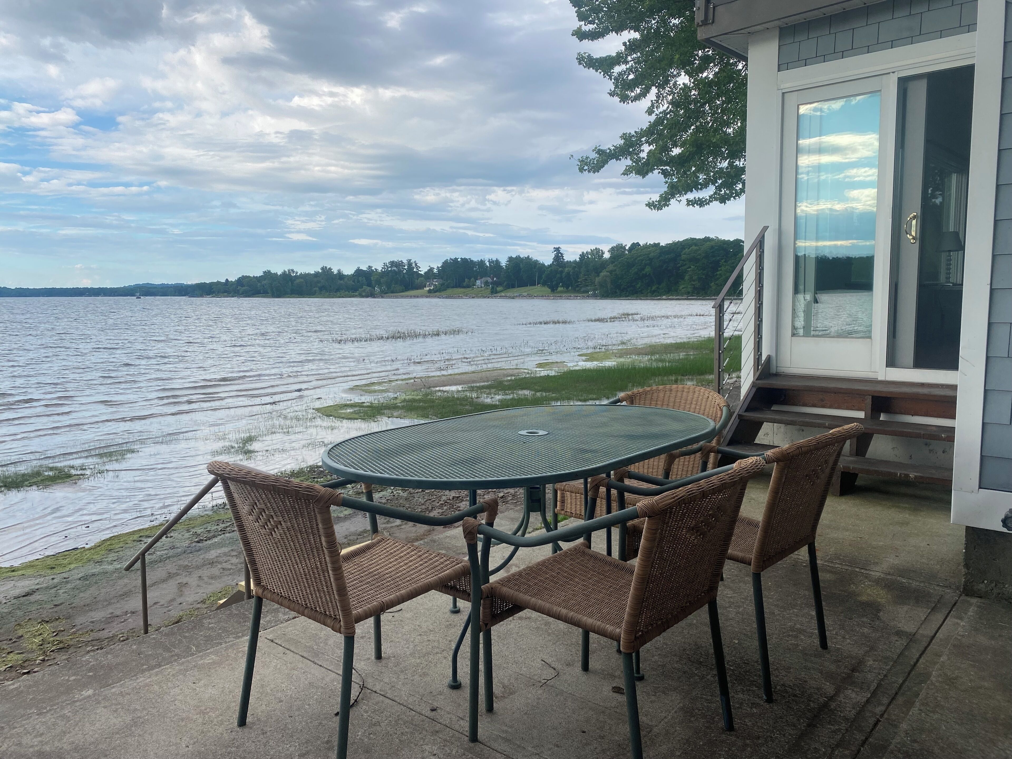 A patio with wicker chairs and a table overlooking a lake, with a house in the background.
