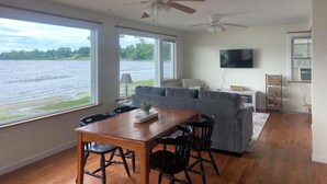 Ett rymligt vardagsrum med ett stort fönster med utsikt över en sjö, en matplats med ett träbord och stolar, en grå soffa och takfläktar.