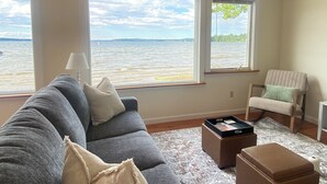 Ett vardagsrum med en grå soffa, en fåtölj, ett soffbord och utsikt över en vattenyta genom stora fönster.