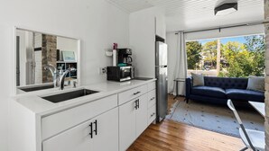 Een moderne keuken met witte kasten, een spoelbak en een koffieapparaat. In de aangrenzende woonruimte staat een donkerblauwe bank en een kleine tafel.